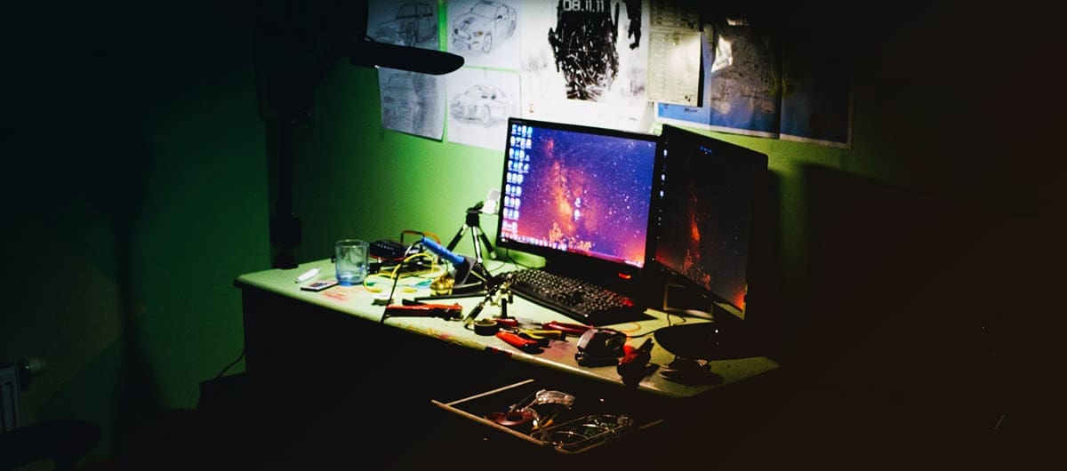 Cluttered desk with two lit monitors and posters hanging on the wall in a dark room with green walls