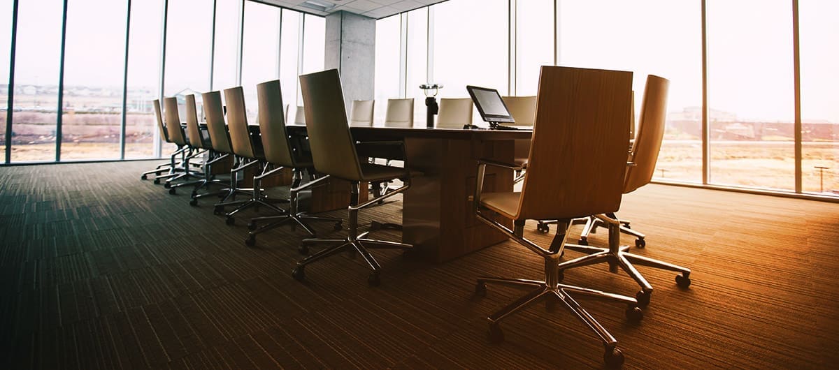 A long, 18-person office table in the middle of a conference room with floor-to-ceiling windows and light beaming through