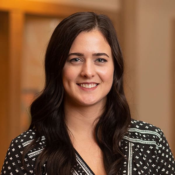 Headshot of a smiling young woman with long, brunette hair dressed in corporate attire in front of a blurred background