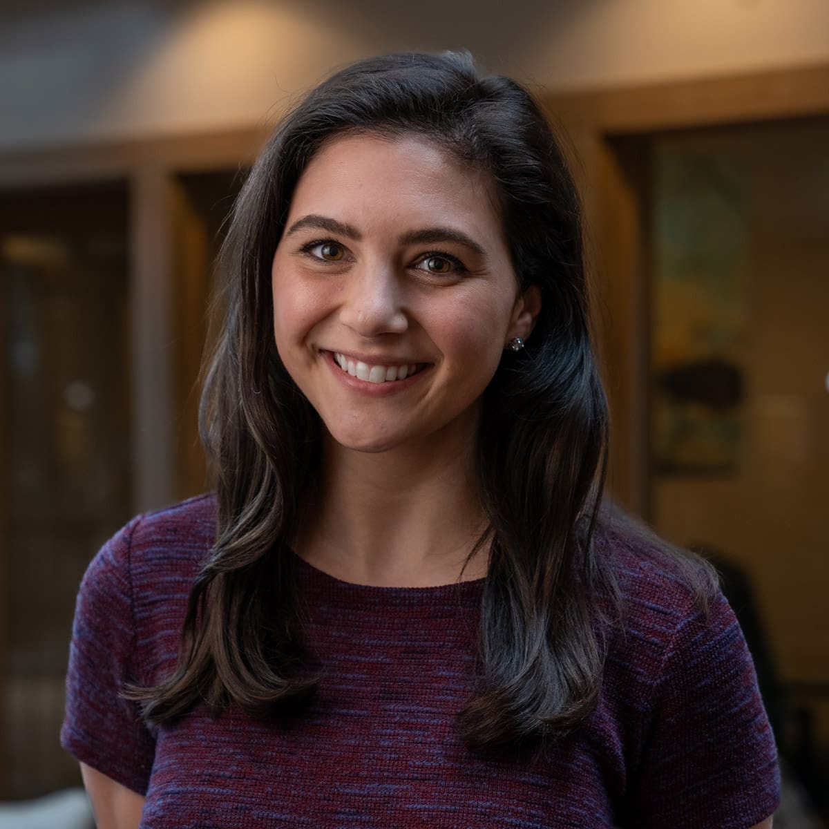 Headshot of a smiling young woman with dark brunette hair dressed in corporate attire in front of a blurred background